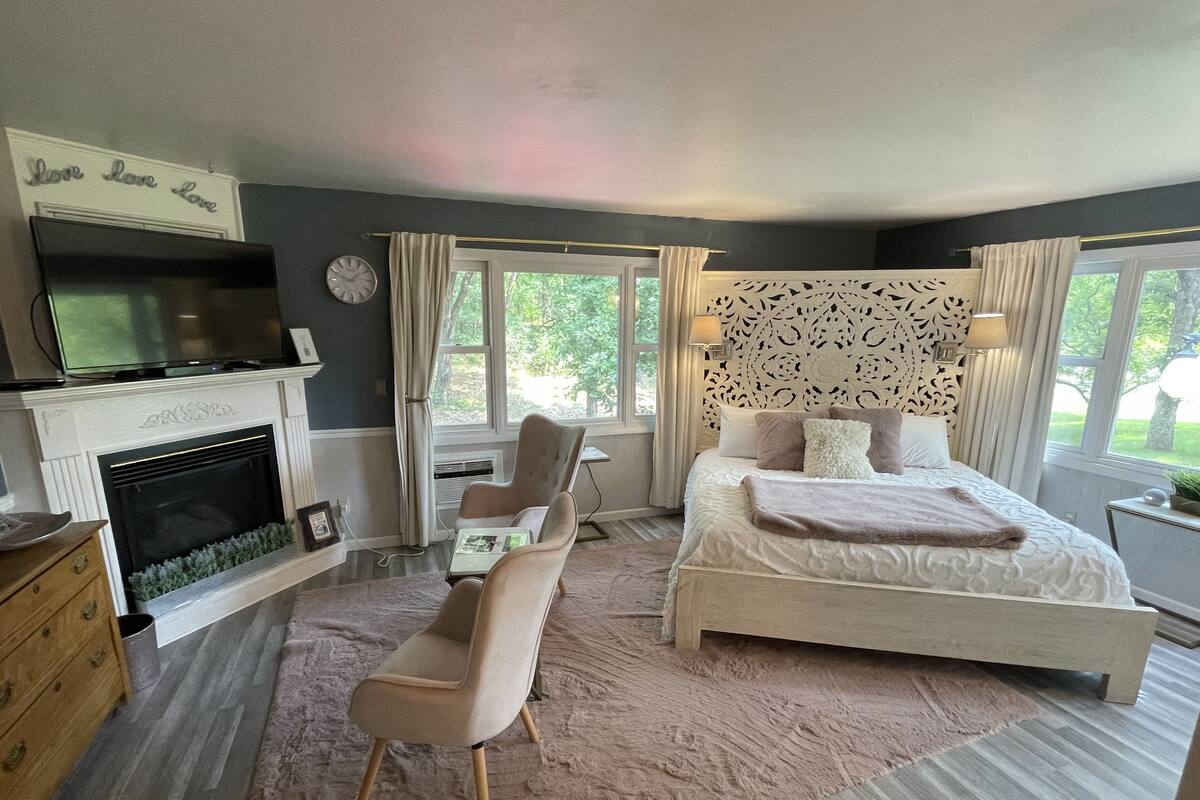 A bedroom with a bed, a chair, a desk, a fireplace, and a television.