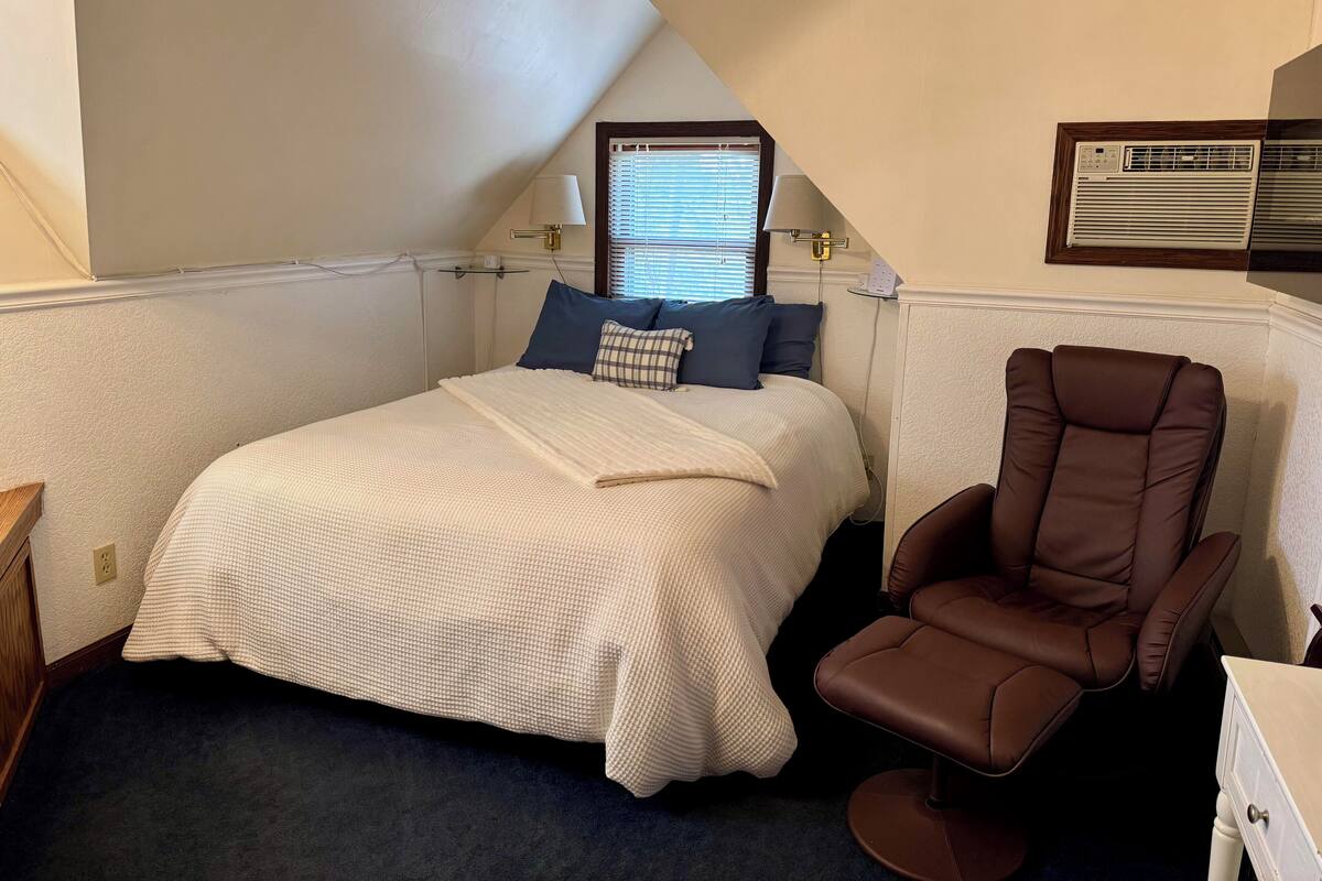 A bedroom with a bed, a brown leather chair, and a window with blinds.