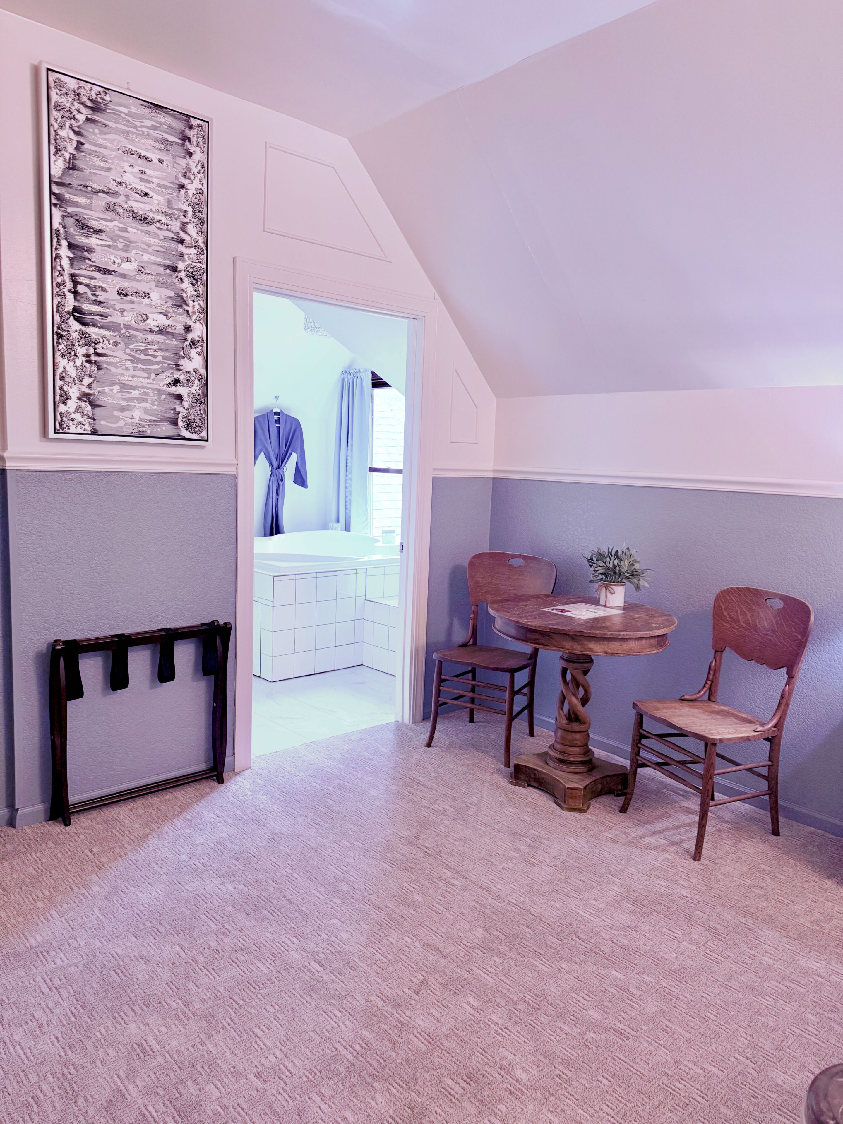 Una habitación con techo inclinado, una mesa de madera con sillas, un perchero y un cuadro abstracto grande.
