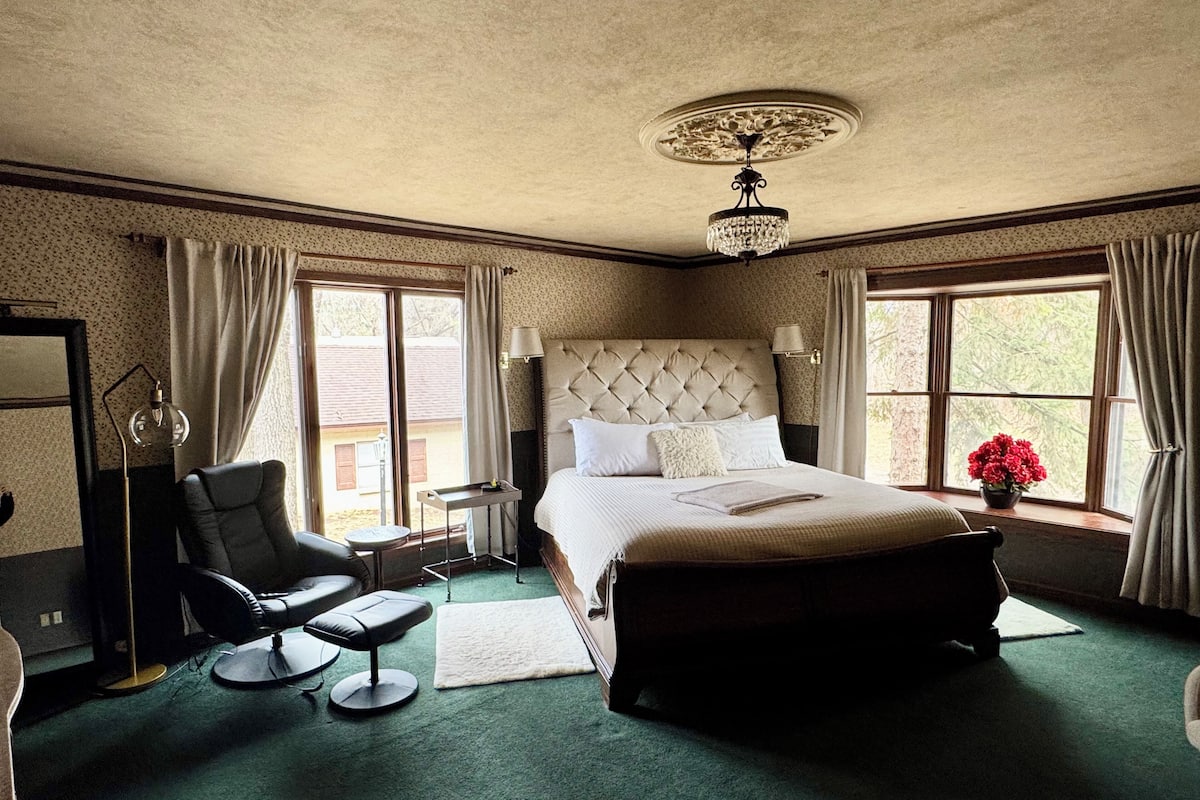 A spacious bedroom with a large bed, a chair, a small table, and a window with curtains.