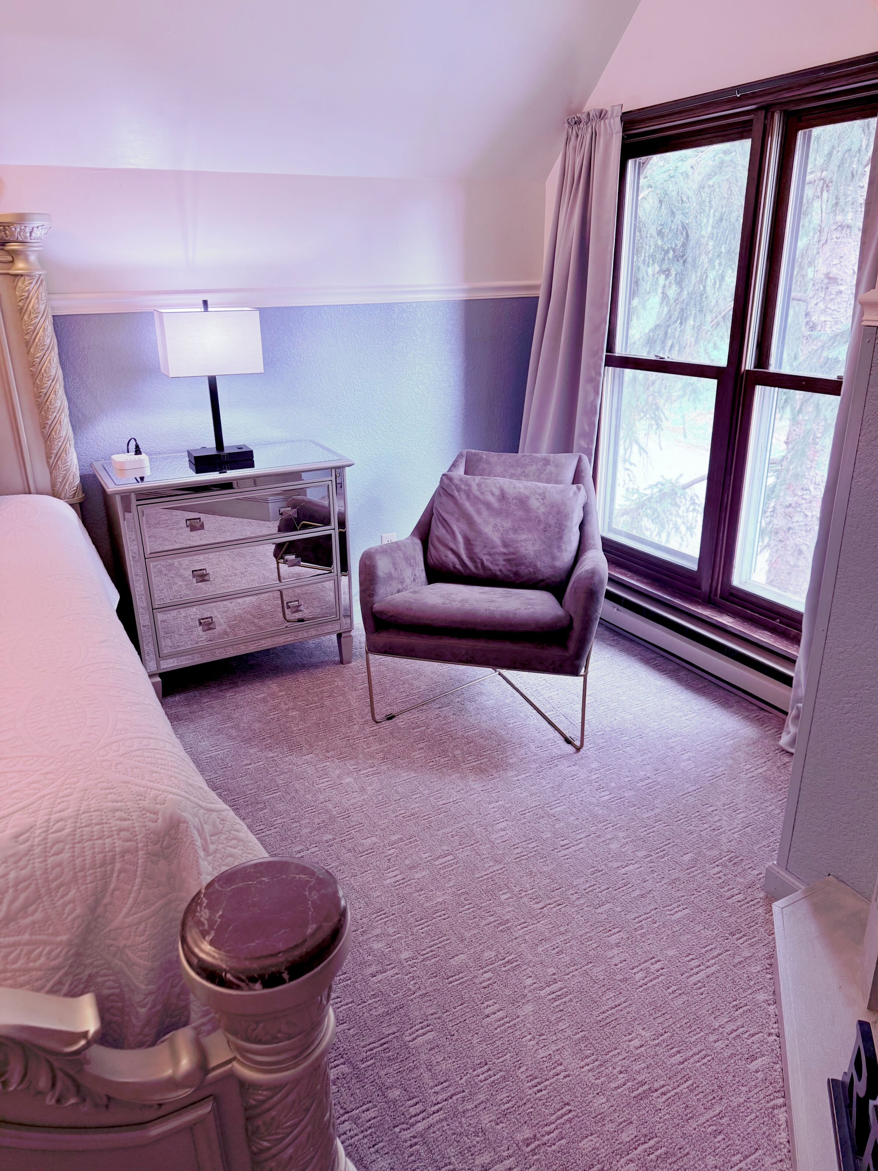 A hotel room with a bed, a chair, a nightstand, a lamp, and a large window.