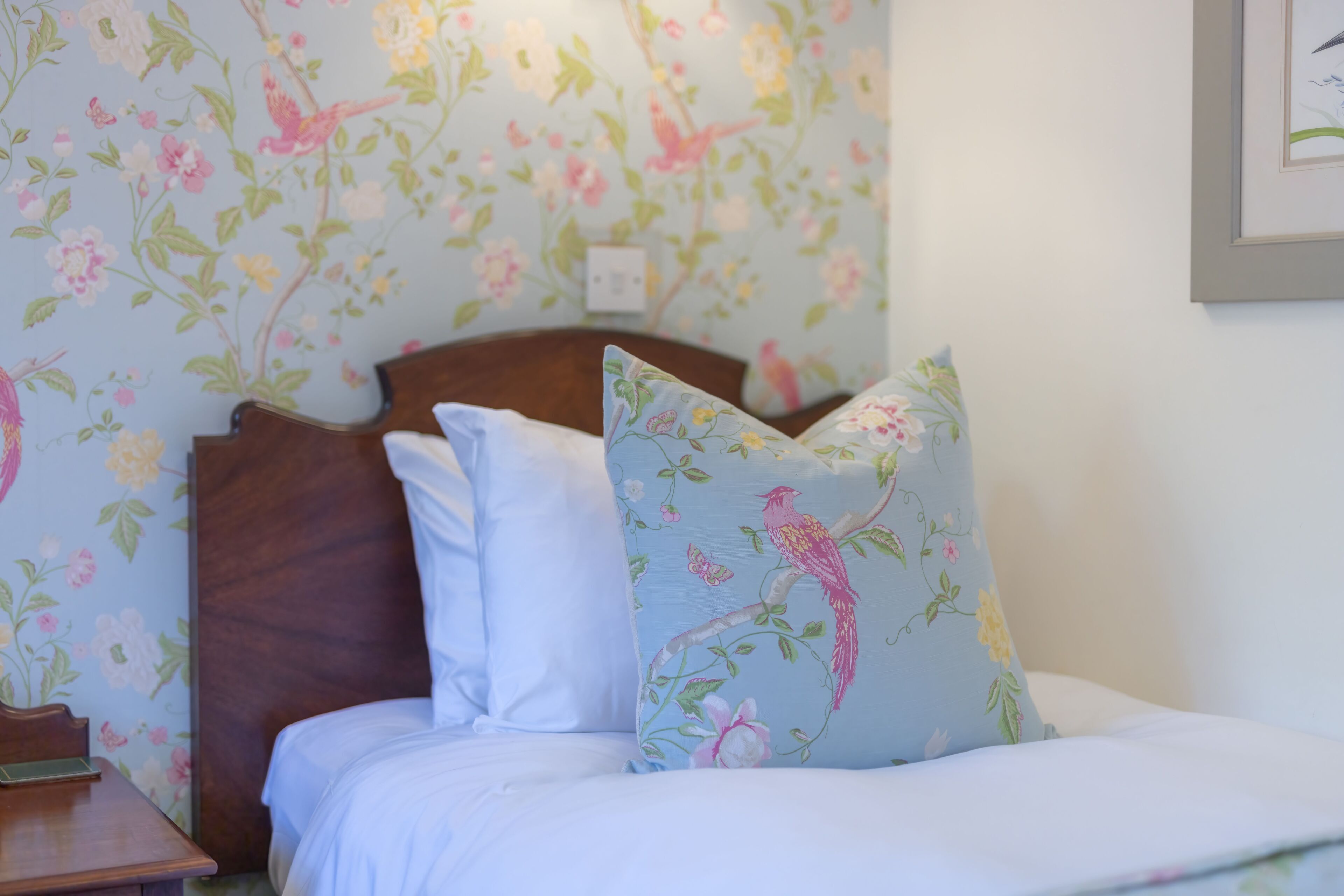 Un dormitorio con papel tapiz floral, cabecera de madera y almohadones decorativos.