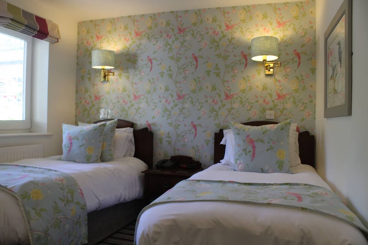 A hotel room with two beds, floral wallpaper, bedside lamps, and a window with curtains.