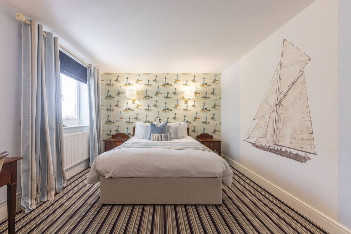 A bedroom with a striped carpet, a bed with white bedding, two bedside tables, and a wall with a nautical wallpaper pattern.