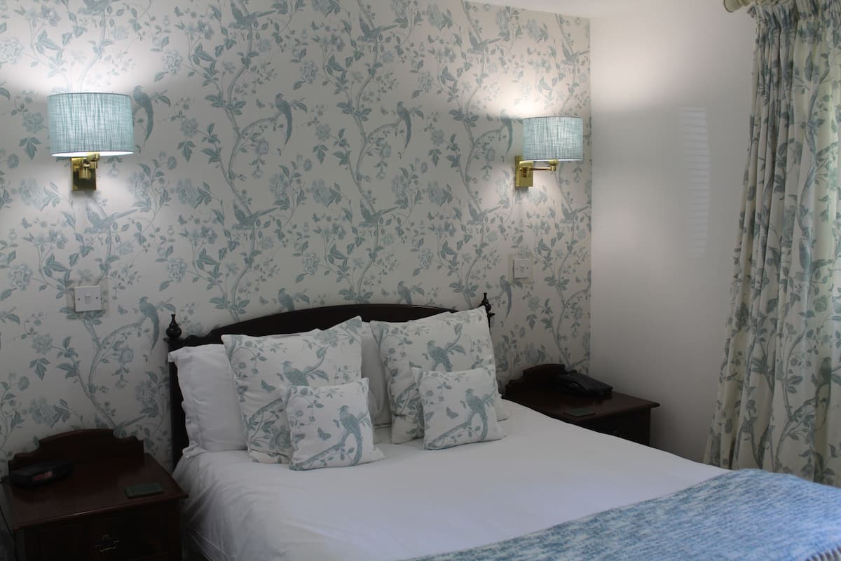 A bedroom with floral wallpaper, a bed with white bedding and matching pillows, two bedside lamps, and a nightstand with a phone and a book.
