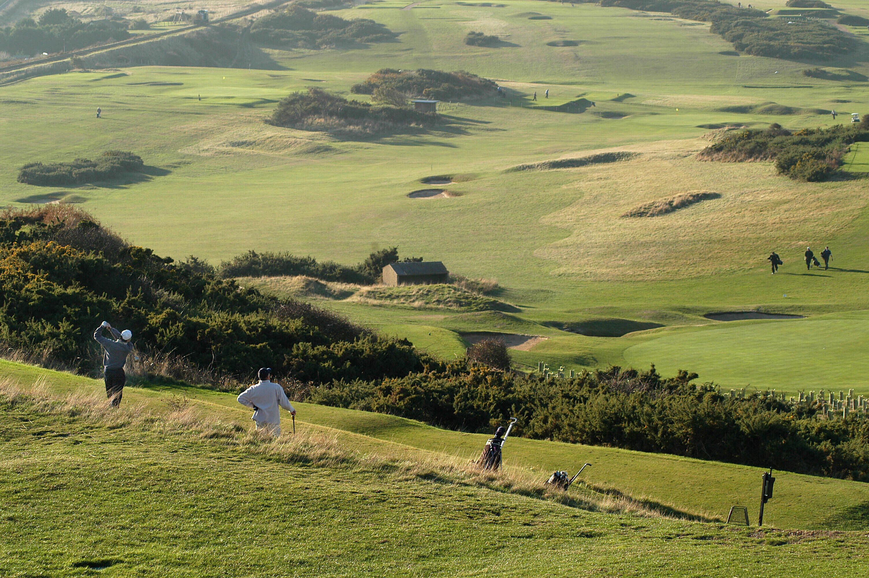 A golf course with rolling hills, bunkers, and a distant water hazard.