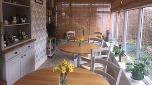 Un comedor con una mesa redonda de madera, sillas blancas y un jarrón con flores amarillas. Se observa una estantería con diversos objetos y una ventana que ofrece vistas al exterior.