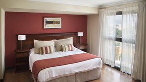 Habitación de hotel con una cama grande, cabecero de madera, dos mesitas de noche con lámparas, una pared de acento roja y vistas a través de cortinas translúcidas.