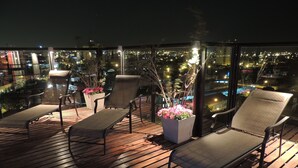 Una terraza en la azotea con suelo de madera, dos sillones de descanso y una maceta con flores rosadas.