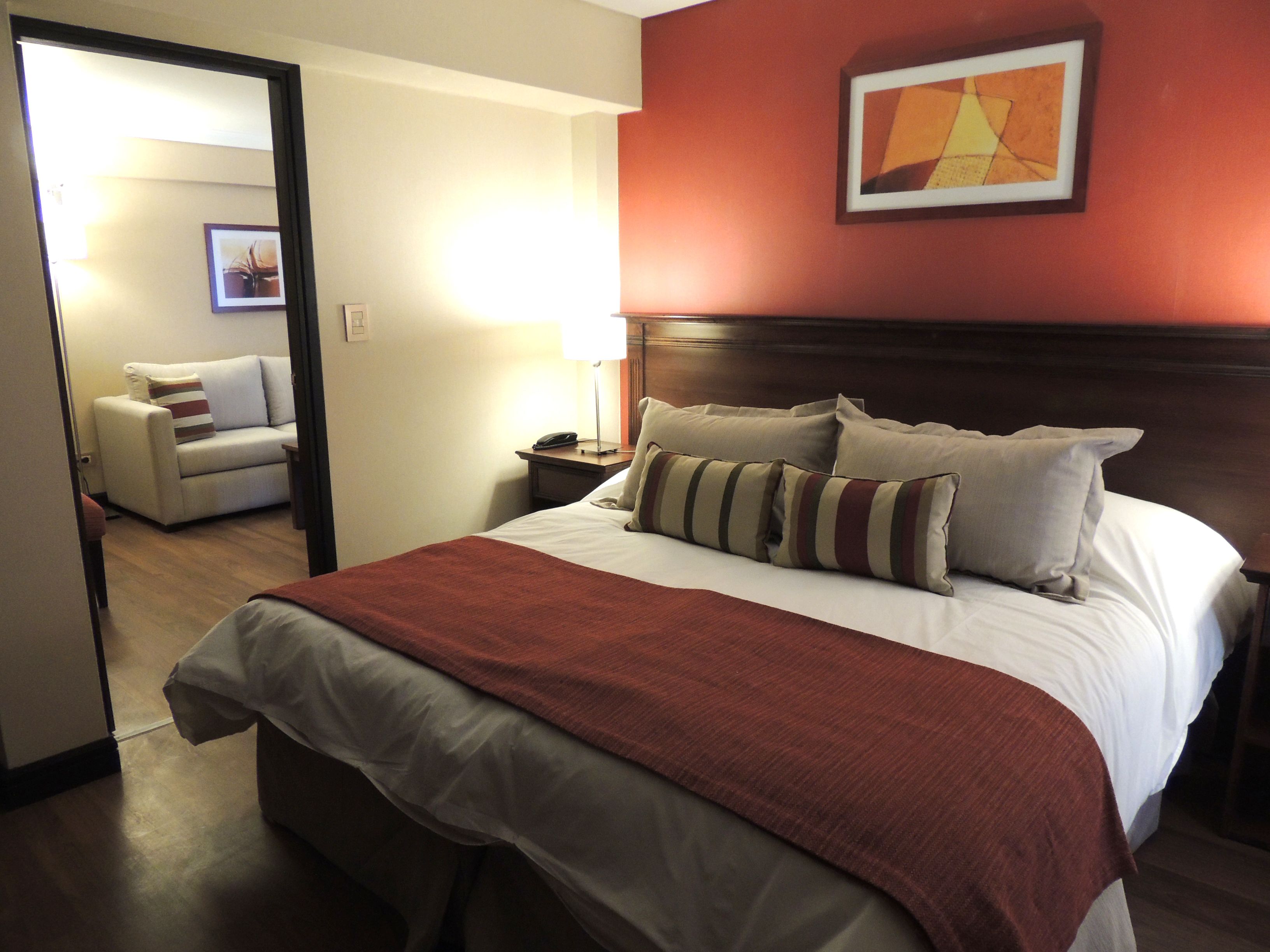 Una camera d'albergo con un letto grande, un comodino, una lampada, una sedia e un quadro appeso al muro.