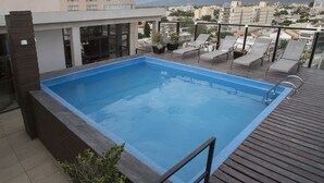 Una piscina en la azotea con terraza de madera y sillones de descanso, rodeada de edificios y montañas al fondo.
