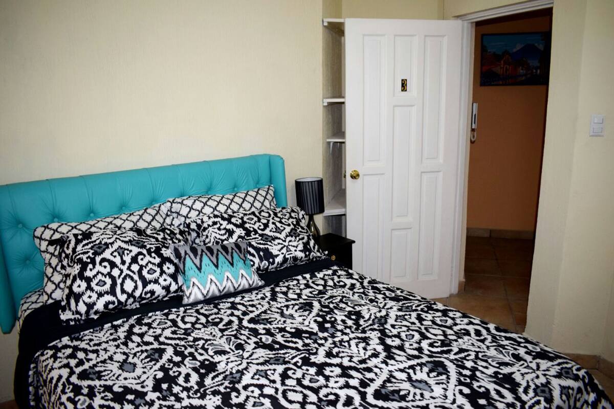 Een slaapkamer met een bed, een blauwgroen hoofdeinde, beddengoed met een patroon, een witte deur en een klein plankje met een lamp.