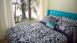 Une chambre à coucher avec une couvre-lit à motifs, une tête de lit recouverte d’un tissu turquoise et une vue sur les arbres par la fenêtre.