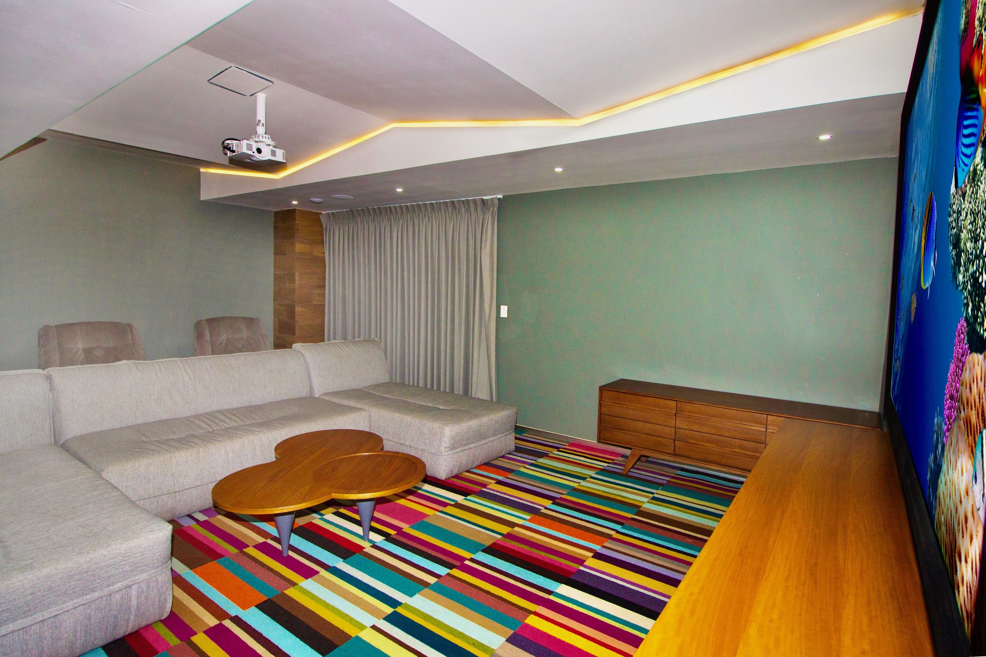 A modern living room with a sectional sofa, a wooden coffee table, a colorful striped rug, a television, and a ceiling projector.