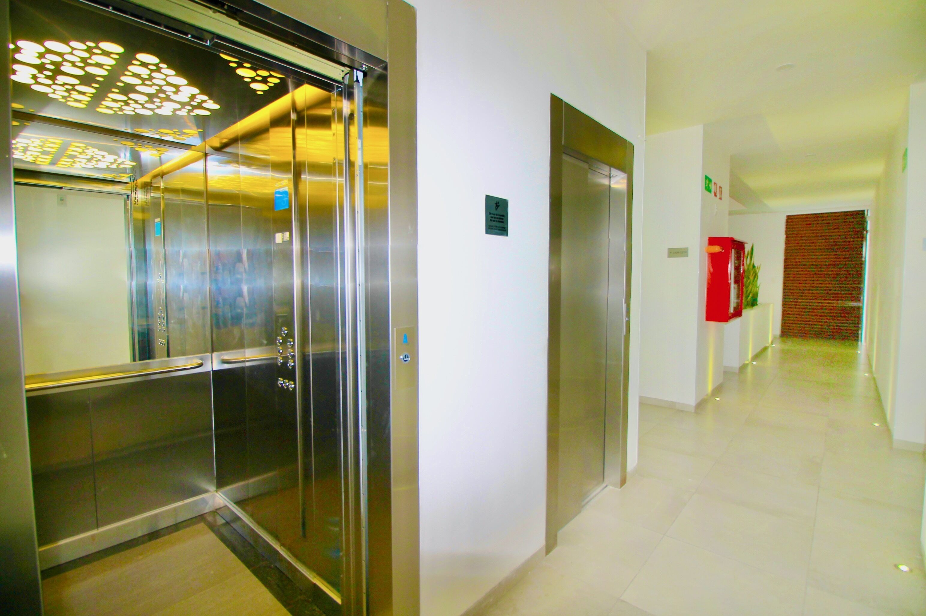 A modern elevator with a metallic finish, a clean hallway with a red object on the wall, and a patterned door at the end.