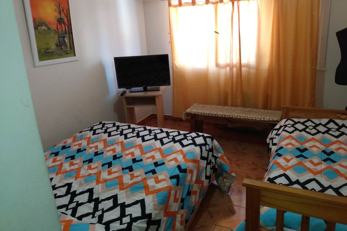 Ein Zimmer mit zwei Betten, einem Fernseher auf einem Schrank, einer Bank und einem Fenster mit Vorhängen.