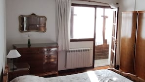 Quarto com cama grande, penteadeira de madeira, mesa de cabeceira com abajur, janela com cortinas e radiador.
