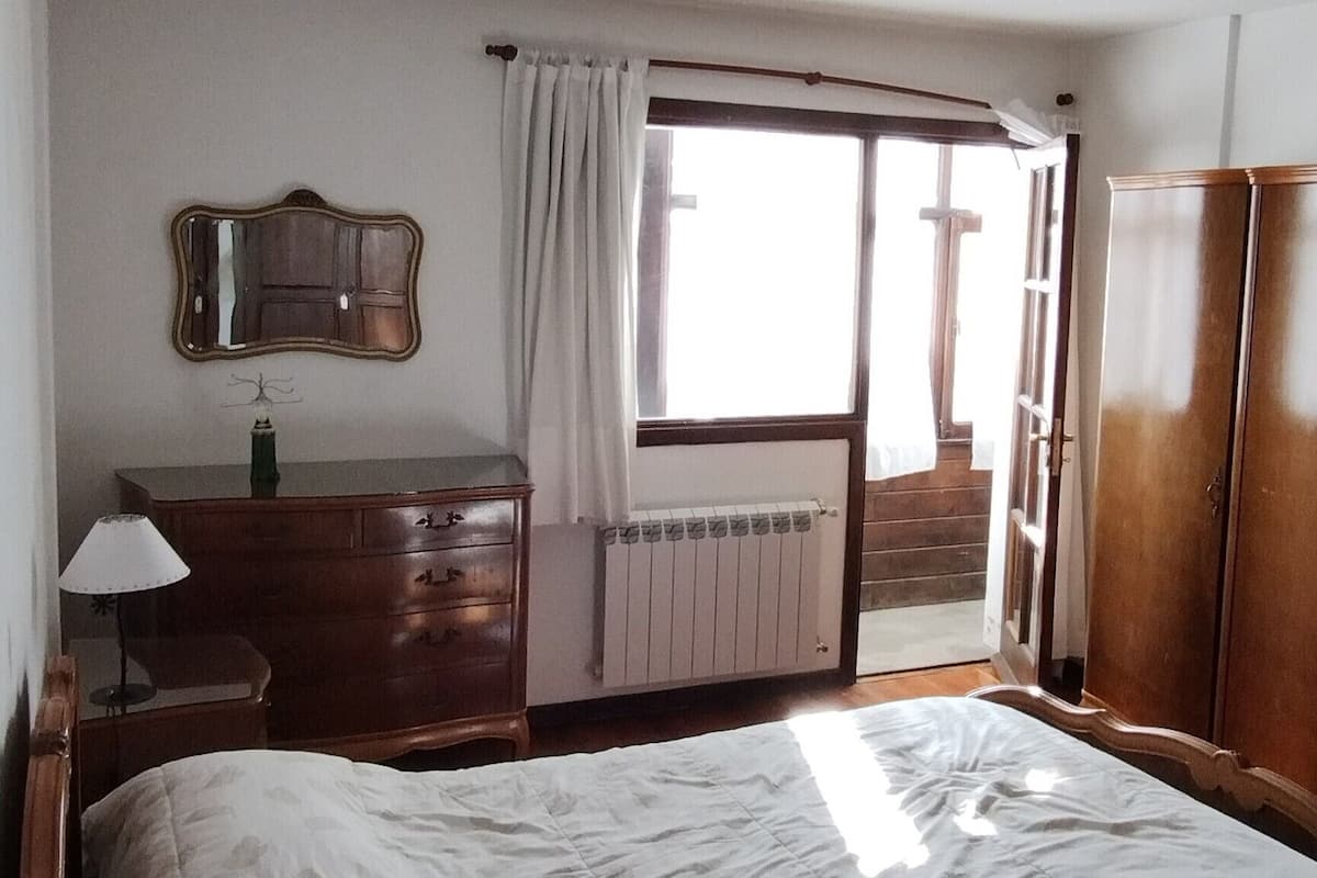 Ein Schlafzimmer mit einem großen Bett, einer hölzernen Kommode, einem Nachttisch mit Lampe, einem Fenster mit Vorhängen und einem Heizkörper.