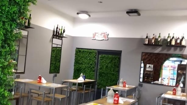 Interior de restaurante com várias mesas e cadeiras, uma parede verde e estantes com garrafas.