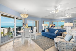 A living room with a blue sofa, white chairs, a dining table, and a view of the ocean through large windows.