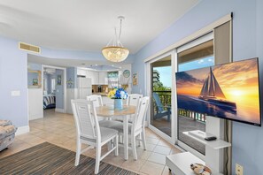 A modern living room with a dining area, a TV mounted on the wall, and a balcony with a view of the sunset.