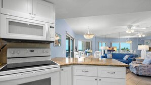 A kitchen with white cabinets, a microwave, and an oven. There is a dining area with a blue couch and a chandelier.