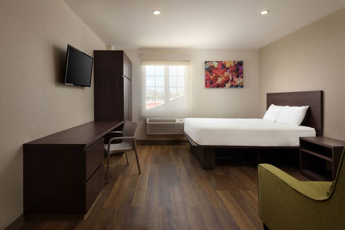 A hotel room with a bed, desk, chair, TV, window, and artwork.