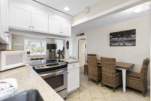 A kitchen with white cabinets, a microwave, stove, and a dining area with wicker chairs and a table. - 🌊 Gulf Winds 2BR Condo — Gulf Views & Steps to the Sand (ST PETE BEACH)