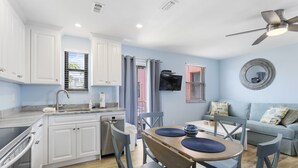 A modern kitchen with white cabinets, a dining area with a wooden table and chairs, a ceiling fan, and a light blue wall.