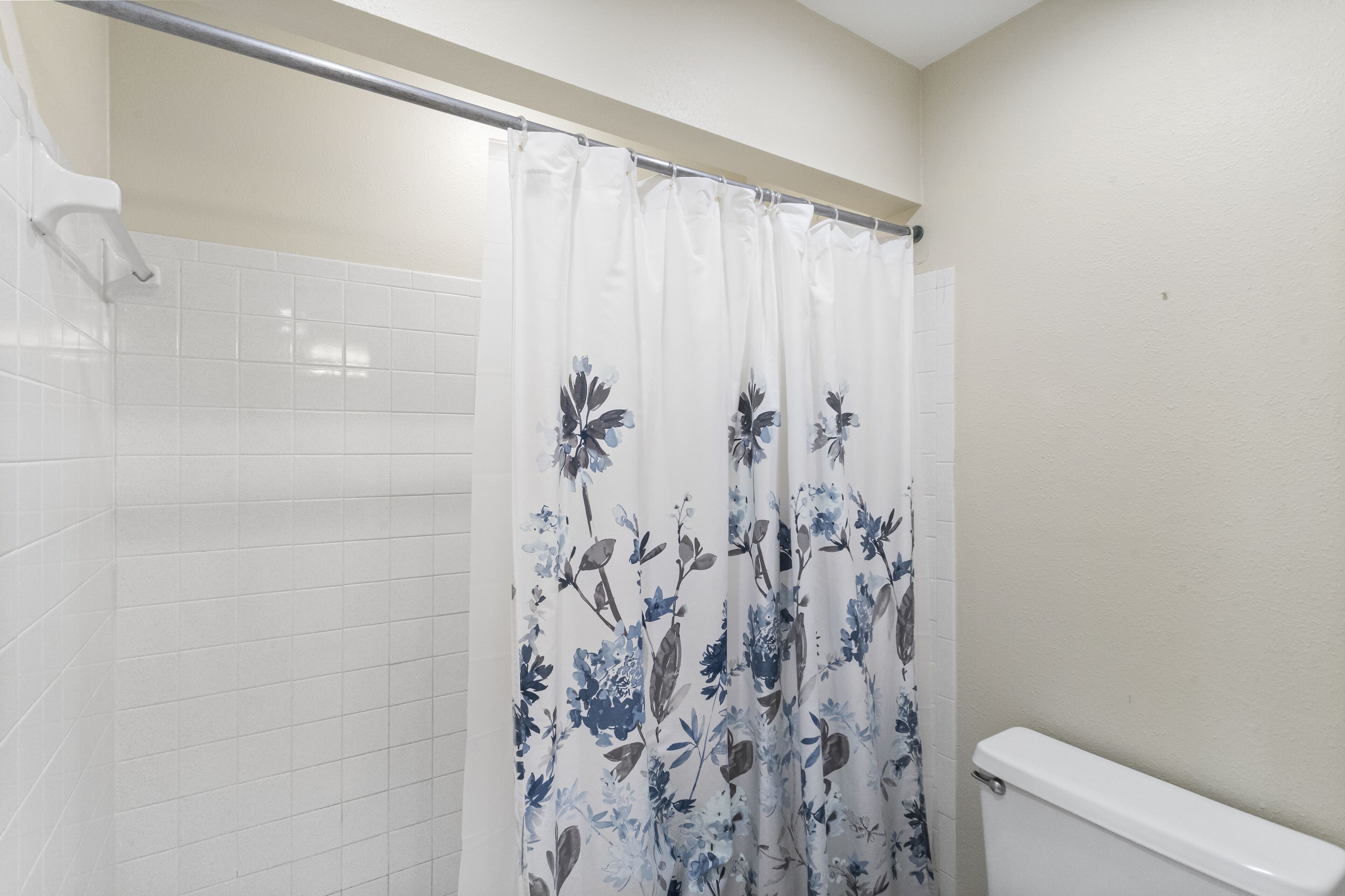 Un baño con paredes de azulejos blancos, una cortina de ducha con estampado floral y un inodoro blanco.