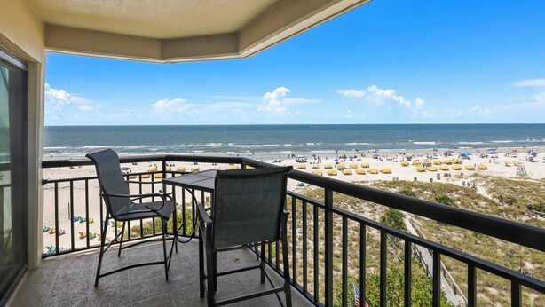 Ein Balkon mit zwei Stühlen und einem Tisch mit Blick auf einen Strand mit Sonnenschirmen und Menschen.