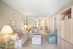 A living room with patterned sofas, a glass coffee table, and a white ottoman. There are lamps on side tables, a dining area with a table and chairs, and a bookshelf.