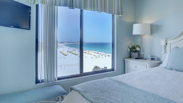 Une chambre d’hôtel dotée d’une grande fenêtre donnant sur la plage, d’un lit, d’une table de chevet avec une lampe et d’un téléviseur fixé au mur.