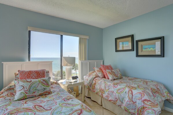A bedroom with two beds, a window with a view of the sea, and two framed pictures on the wall.