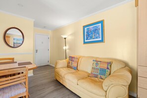 A room with a beige sofa, a wooden dining table, a mirror, a floor lamp, and a framed picture on the wall. - Gulf Winds 508 | Affordable Studio on Upham Beach (ST PETE BEACH)