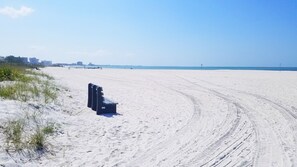 Una playa arenosa con un banco y marcas de neumáticos, cielo despejado y edificios a lo lejos.