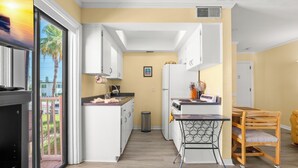A modern kitchen with white cabinets, a small island, a trash bin, and a dining area with a table and chairs. - Gulf Winds 508 | Affordable Studio on Upham Beach (ST PETE BEACH)