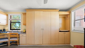 A room with a wooden wardrobe, a desk with a chair, a window with blinds, and a red cushioned bench. - Gulf Winds 508 | Affordable Studio on Upham Beach (ST PETE BEACH)