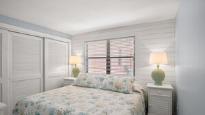 Un dormitorio con cama, dos mesitas de noche con lámparas, una ventana con persianas y una puerta con contraventanas.