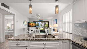 Cocina con armarios blancos, encimeras de granito y electrodomésticos de acero inoxidable. Cuenta con un ventilador de techo, luces colgantes y vistas al exterior a través de una ventana.