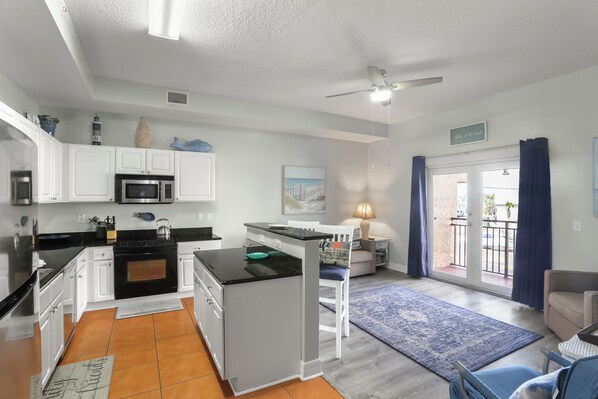 Una cocina moderna con armarios blancos, encimera negra y una isla central. Hay una zona de comedor con un tapete azul y una silla blanca.