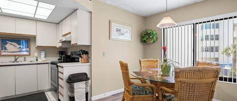 A modern kitchen with white cabinets, a dining area with wicker chairs, and a large window with blinds.