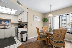 A modern kitchen with white cabinets, a dining area with wicker chairs, and a large window with blinds.
