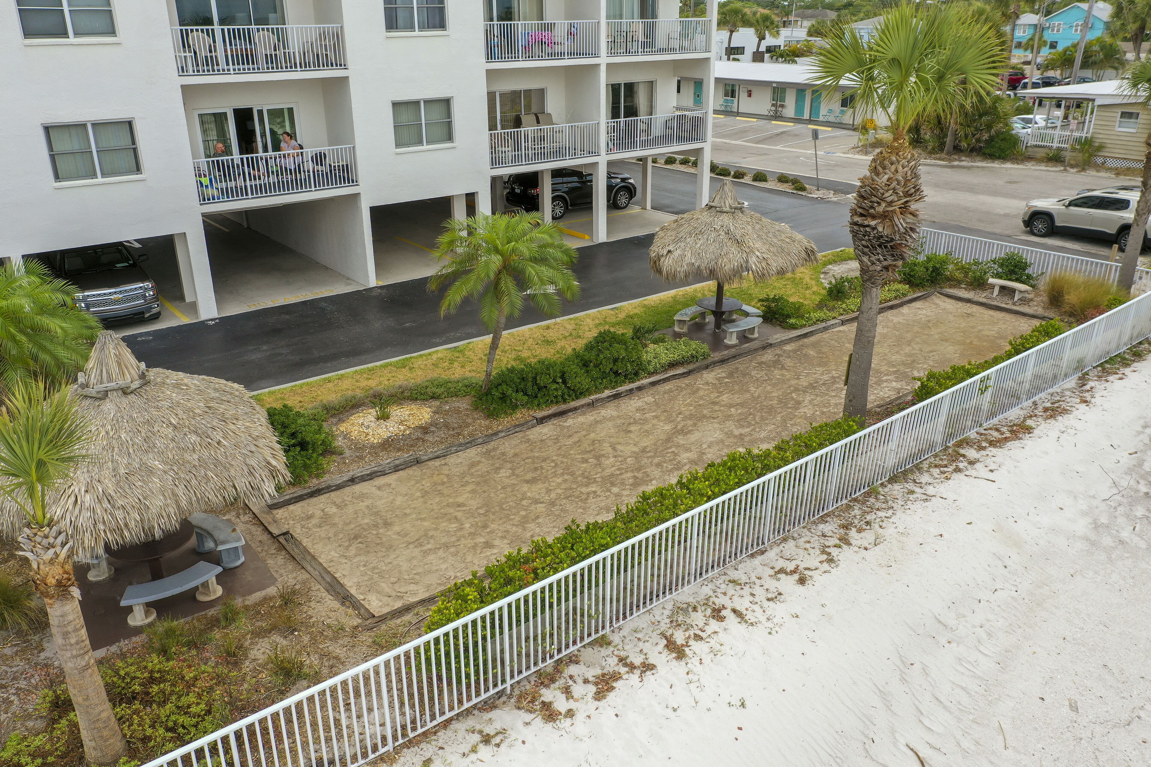 A multi-story hotel with balconies, a parking area, and a landscaped garden with palm trees and a thatched-roof structure.