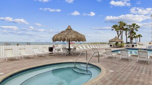 A pool area with lounge chairs, a thatched umbrella, and palm trees. - Sea Breeze 301 | Corner Unit with Dual Balconies & Sweeping Gulf-to-Bay Views (Madeira Beach)