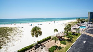 A beach view with a pool area, palm trees, and beach umbrellas. - Sea Breeze 301 | Corner Unit with Dual Balconies & Sweeping Gulf-to-Bay Views (Madeira Beach)