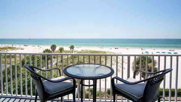 A balcony with two chairs and a table overlooking a beach with umbrellas and people. - Sea Breeze 301 | Corner Unit with Dual Balconies & Sweeping Gulf-to-Bay Views (Madeira Beach)