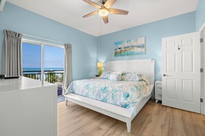 Une chambre à coucher avec un lit, un ventilateur de plafond, une fenêtre avec des rideaux, un tableau au mur et une porte.