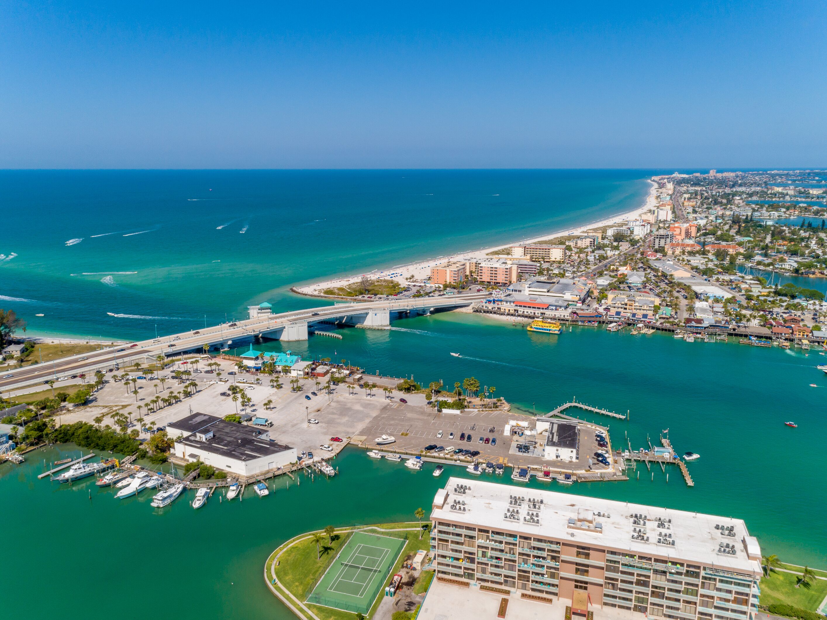 An aerial view of a coastal city with a marina, a bridge, and a large hotel with multiple floors and balconies.