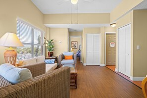 A spacious living area with wicker furniture, a glass coffee table, and a view of the outdoors through large windows.